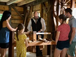 A family looks on as a cabinetmaker in 1830s costume works a treadle wheel