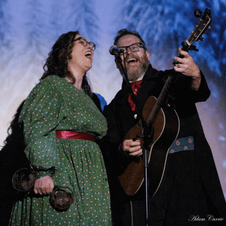 Two people, one of which is playing a guitar, dressed in 1830s clothes, sing lively music