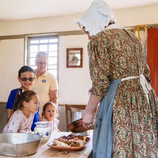 A costumed historian in 1830s dress shows a plate of hearth cooked food to kids and an adult