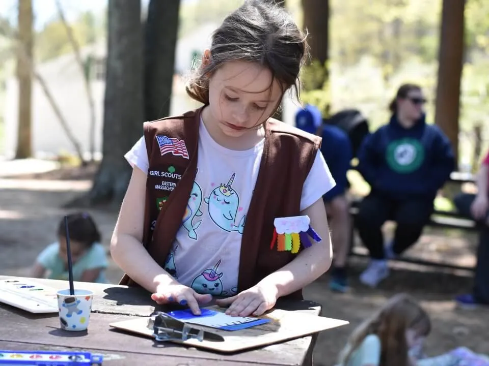 A young girl scout (wearing a brownie uniform) paints on a picnic table