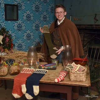 A person in 1830s attire holds up a hand-knit stocking and stands by a table with Christmas stckings, wooden shoes, and a candle