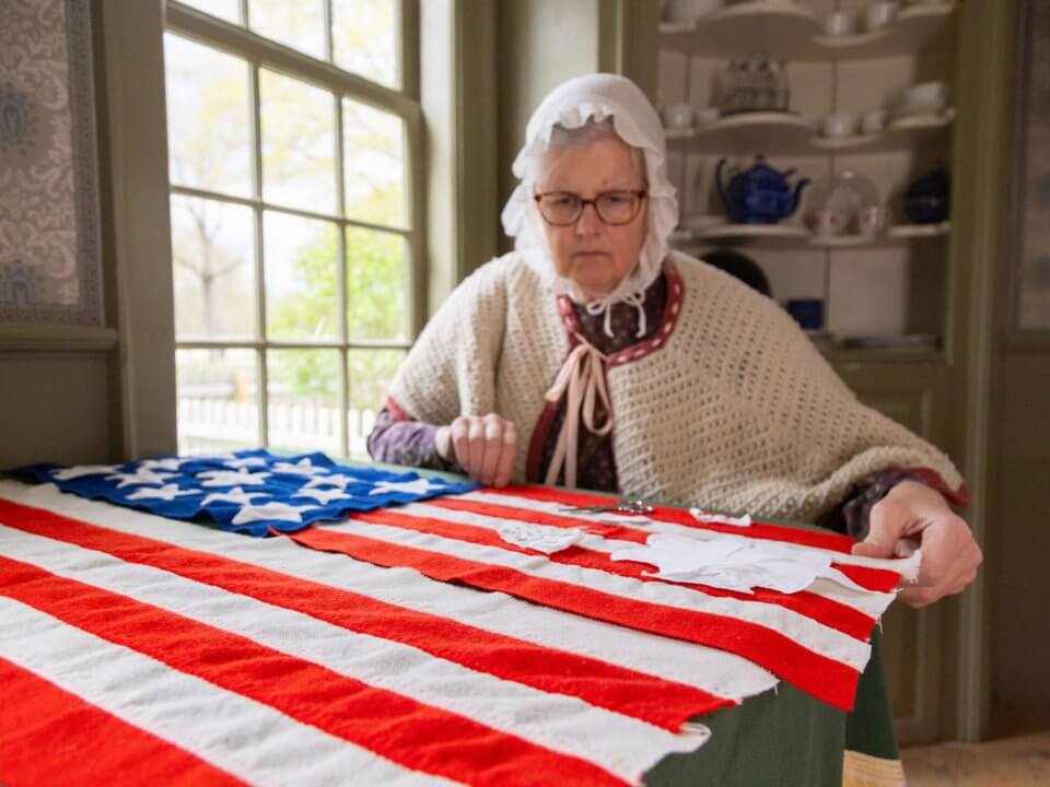A woman in 1830s attire hand-sews an Amercian Flag