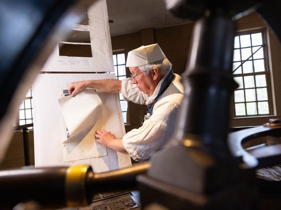 A man in 1830s attire works a 19th-century printing press