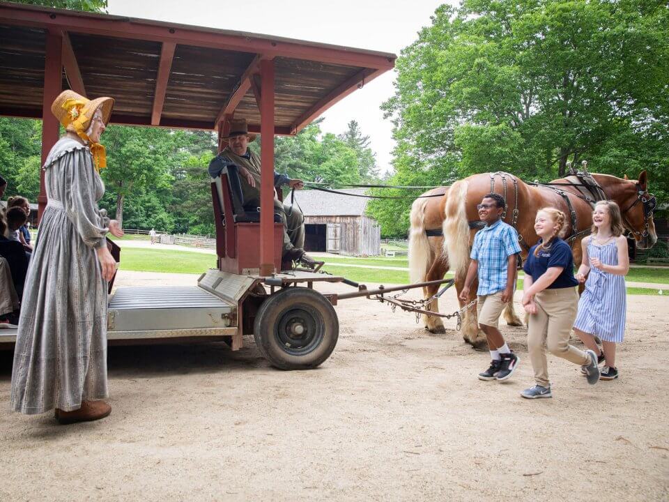 Three kids approach a horse drawn wagon driven by a man in 1830s clothing