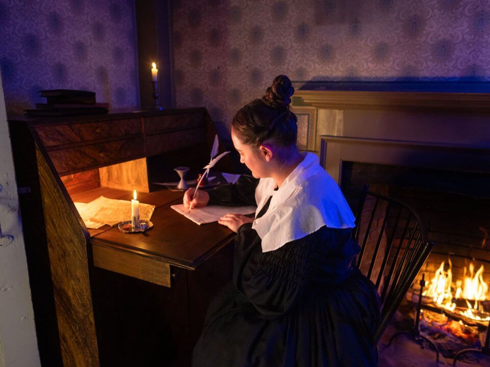 A woman in 1830s attire sits and writes with a quill pen by candlelight